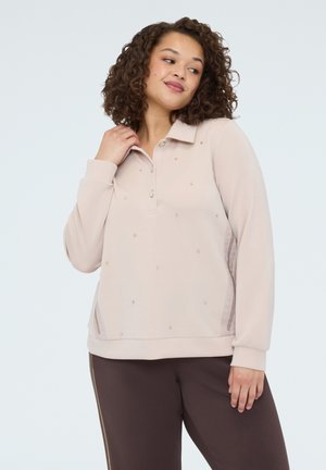 Femme aux cheveux bouclés portant un pull beige à manches longues avec de petites ornements et un pantalon marron foncé sur un fond uni.
