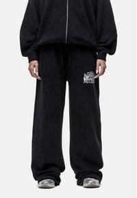 Zwarte pullover hoodie met ritssluiting aan de voorkant en grote zakken aan de voorkant, gecombineerd met losse zwarte joggingbroeken met een geprinte ontwerp op het been.