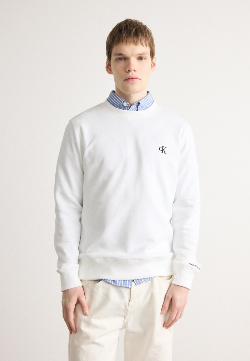 Sudadera blanca con puños y dobladillo acanalados, que presenta un logo negro de "CK" en el pecho, superpuesta a una camisa a rayas azul clara.
