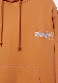Sweat à capuche orange rouille avec poche avant, cordons de serrage et texte brodé blanc "design studios air nuances" accompagné de trois étoiles sur la poitrine.
