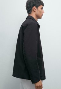 Black jacket with a structured collar and side pockets, featuring a straight cut and smooth texture, worn over a white shirt.