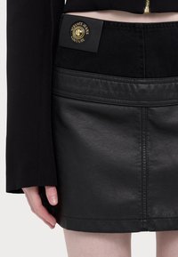 Svart läder mini kjol med texturerade paneler och midjeband i denim. Har en guldfärgad logotagg och synliga sömnadsdetaljer.