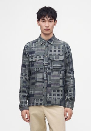 Jeune homme portant une chemise à manches longues boutonnée avec différents motifs géométriques gris et noirs et un pantalon beige sur un fond uni.
