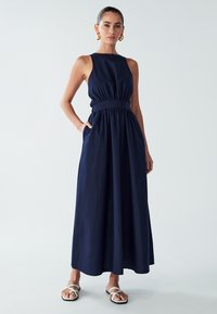 Kvinna står i en ärmlös marinblå maxiklänning med insydd midja och vita, remmade platta sandaler, mot en enkel ljus bakgrund.