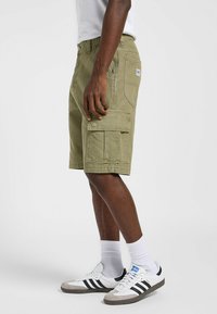 Shorts cargo vert olive avec plusieurs poches, en tissu coton et à coupe décontractée. Associés à des baskets blanches et grises, et des chaussettes blanches.