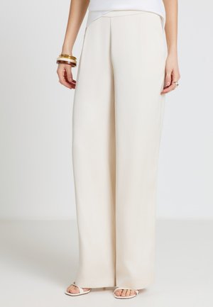 Femme portant un pantalon crème taille haute à jambes larges, associé à des sandales blanches, un haut blanc et des bracelets dorés et bruns au poignet gauche.