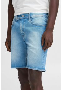 Shorts en denim bleu clair avec une coupe droite, cinq poches, et une fermeture à bouton métallique et zip. Coutures contrastantes le long des coutures.