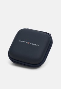 Étui rigide noir Tommy Hilfiger avec fermeture éclair, texture lisse, forme carrée, présentant le logo de la marque en relief à l'avant.