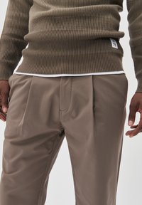 Hnědý žebrovaný pletený svetr kombinovaný s světlými hnědými kalhotami s puky a bočními kapsami, umístěný na jednolitém pozadí.