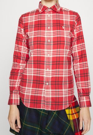 Femme portant une chemise à carreaux rouge et blanc à boutons, manches longues et col, associée à une jupe à carreaux foncés avec des accents jaunes.