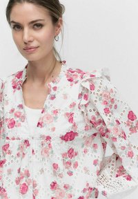 Hvid blomstret bluse med lyserøde og grå roser, blonde detaljer på ærmerne, flæser og en V-hals. Stoffet fremstår let og tekstureret.