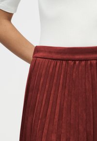 Κόκκινη φούστα με πιέτες, λεία, υφή και φαρδιά μέση. Το φόντο είναι απλό, δίνοντας έμφαση στο σχέδιο της φούστας.