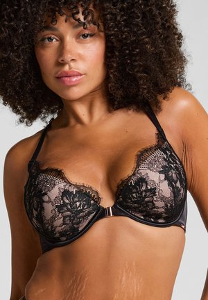 Mujer con cabello rizado que lleva un sujetador de encaje negro con patrones florales, mostrando su torso superior y con una expresión facial neutra.