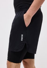 Άνδρας με μαύρο στρωματοποιημένο αθλητικό σορτς με το λογότυπο "BORG" στο εξωτερικό μέρος του σορτς, χέρι στην τσέπη, σε απλό ανοιχτό φόντο.