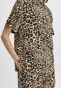 Chemise à manches courtes en motif léopard, fabriquée en tissu doux, avec une fermeture à boutons sur le devant et une coupe décontractée. Palette de couleurs marron et beige.