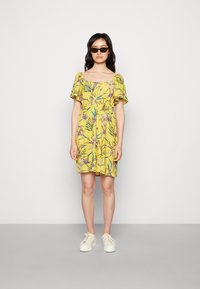 Vestido amarelo floral com mangas curtas e leves, cintura franzida e um corte relaxado. Combinado com ténis brancos e óculos de sol estilosos.