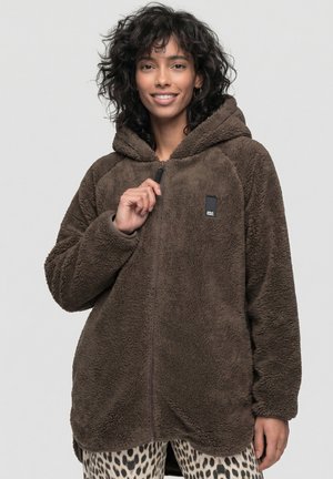 Frau mit lockigem Haar zieht den Reißverschluss einer übergroßen braunen Fleecejacke mit Kapuze hoch, trägt eine Hose mit Leopardenmuster und lächelt vor einfachem Hintergrund.