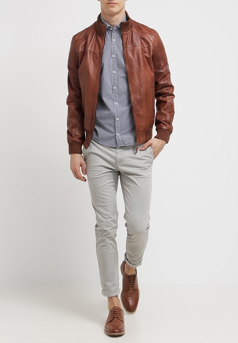 Homme portant une veste en cuir marron, une chemise à carreaux bleue, un pantalon gris clair et des chaussures en cuir marron, debout avec une main dans la poche.