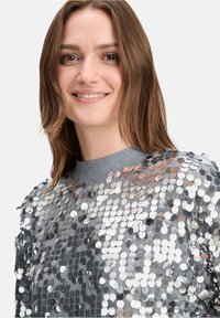 Silberner Paillettenoberteil mit einem grauen Kragen, versehen mit dicht angeordneten metallischen Kreisen über den Stoff für eine reflektierende Textur.