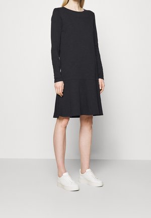 Person, die ein langärmeliges schwarzes Kleid mit ausgestelltem Rock und weißen Plateausneakern trägt, steht vor einfachem Hintergrund.