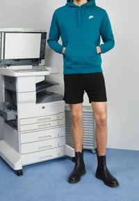 Tyrkysová mikina Nike s předním kapsou, černé síťované šortky a černé kožené kotníkové boty s žlutými akcenty, vedle tiskárny.