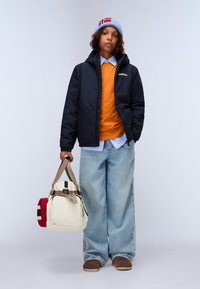 Veste matelassée marine, pull orange, chemise bleu clair, jean large délavé. Porte un sac crème avec des anses marron et des accents rouges.