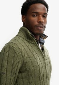 Pull en tricot côtelé vert olive avec un col haut, un motif texturé et une chemise à carreaux superposée visible au col.