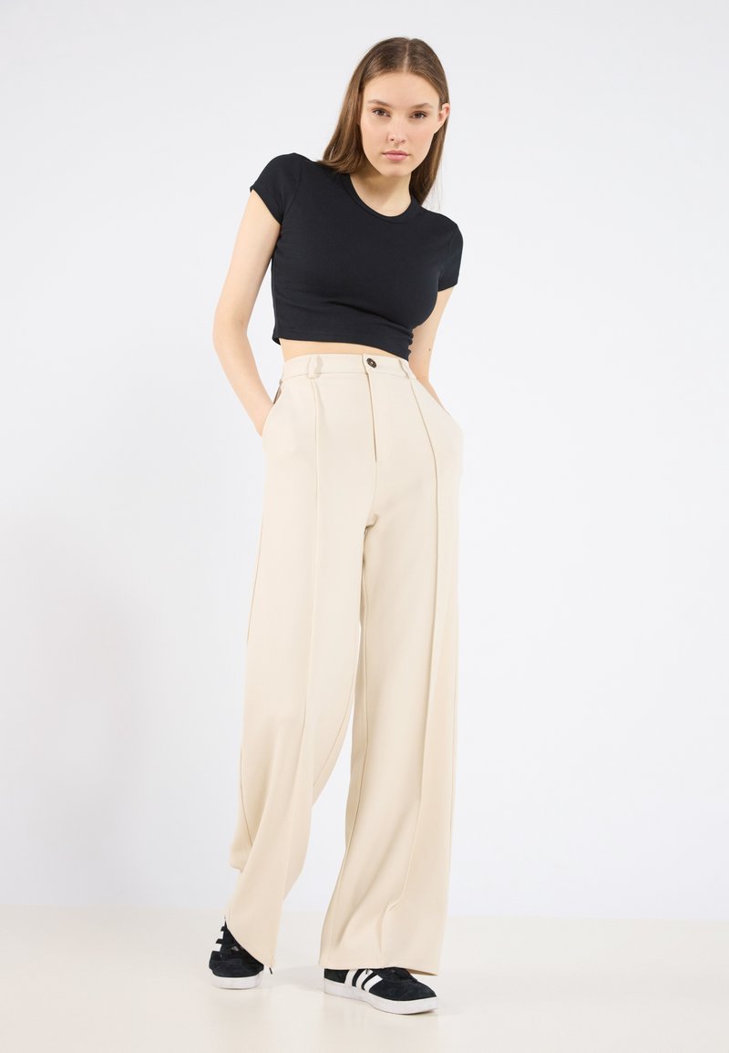 Beige hoge taille brede broek met voorplooien, knoopsluiting en zijzakken, gecombineerd met een zwart cropped t-shirt en zwarte sneakers.