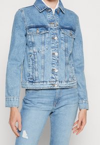 Chaqueta de mezclilla azul claro con un corte clásico, dos bolsillos frontales y cierre de botones. Un sutil desvanecimiento y detalles de costura realzan la textura.