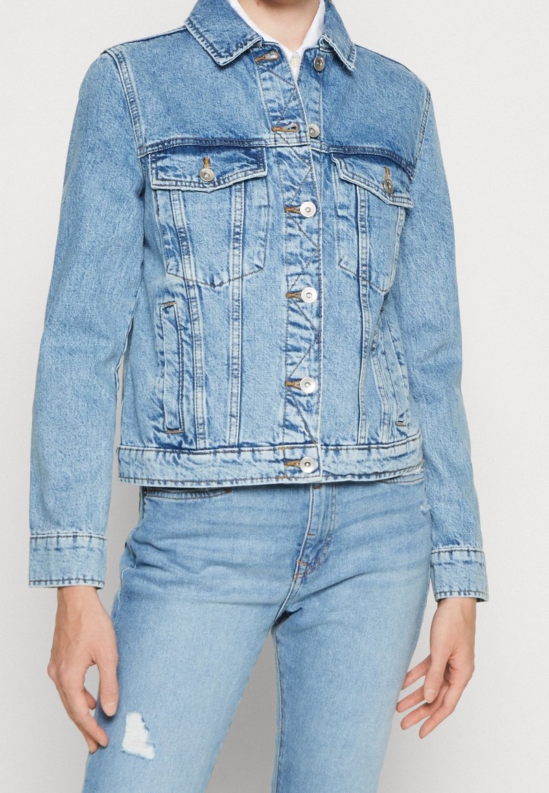 Chaqueta de mezclilla azul claro con un corte clásico, dos bolsillos frontales y cierre de botones. Un sutil desvanecimiento y detalles de costura realzan la textura.