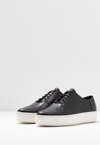 Zapatillas de cuero negro con una textura suave, diseño de perfil bajo, suela de goma blanca y cordones en la parte delantera con seis ojales.