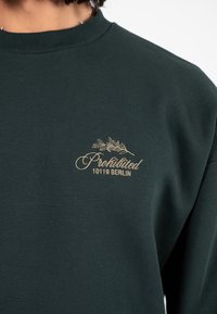 Felpa verde scuro con testo ricamato beige "Proibito 10119 Berlino" e un piccolo ramo d'olivo sopra il testo sul petto.