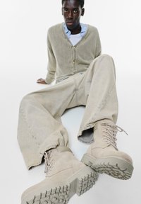 Beige velúr csizmák texturált talppal, oversize világos bézs farmernadrággal párosítva. A modellen világosszürke kardigán van egy kék ing felett.