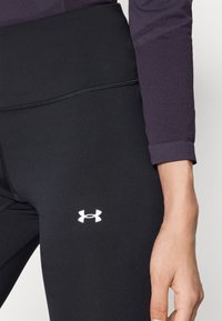 Černé legíny vyrobené z elastického materiálu s hladkou texturou. Na stehně se nachází bílé logo Under Armour a detaily šití.