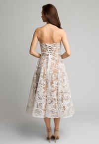 Sukienka bez ramiączek, przezroczysta biała, z kwiatowym koronkowym wykończeniem, dopasowanym gorsetem i rozkloszowanym dołem. Posiada szczegółowe zawiązanie na plecach oraz kremowe szpilki.