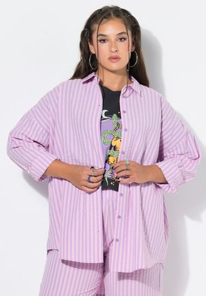 Junge Frau mit langen dunklen Haaren in zwei vorderen Zöpfen trägt Creolen, ein pink-lila gestreiftes Hemd und Hose sowie ein schwarzes grafisches T-Shirt.