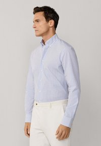 Muškarac u svijetloplavoj kariranoj košulji na kopčanje, uvučenoj u bijele hlače, stoji ispred jednobojne svijetlosive pozadine.