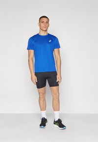 Blå atletisk t-shirt med korta ärmar, kombinerad med svarta kompressionsshorts. Modellen har på sig svarta sneakers med gröna detaljer.