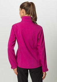 Mujer con coleta castaña, vistiendo una chaqueta magenta y pantalones negros, de pie de espaldas contra un fondo claro y sencillo.