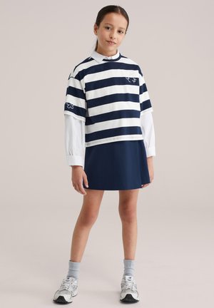 Krátký modrý a bílý proužkovaný top přes bílou košili s dlouhým rukávem, doplněný modrou sukní a šedými ponožkami, s stříbrnými teniskami.