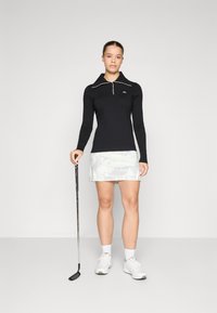 Black long-sleeve polo with a zip collar, featuring white piping. Paired with a light-patterned skort. Golf club held in right hand.