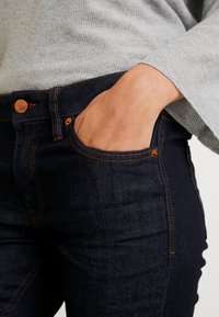 Jeans de mezclilla azul oscuro con costuras naranjas, que presentan un diseño de bolsillo en la parte frontal. El material parece suave con un ligero brillo.