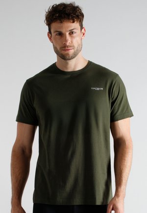Man with short curly hair wearing olive green short-sleeve Tom Tailor t-shirt standing against gray background.