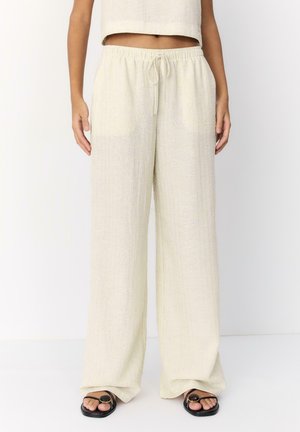 Donna che indossa pantaloni larghi di colore beige chiaro con una texture e una vita con coulisse, abbinati a sandali neri aperti sulle dita, davanti a uno sfondo bianco uniforme.