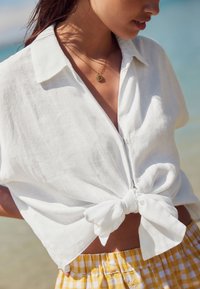 Hvid hørskjorte med korte ærmer, knyttet ved taljen. Kombineret med et gult og hvidt ternet skørt. Enkel gylden halskæde synlig.