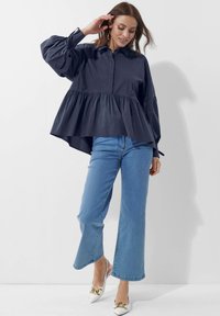Dunkelblauses Hemd mit Kragen und gepufften Ärmeln, gestuftem Design und abgerundetem Saum. Kombiniert mit hellblauen, ausgestellten Jeans und weißen hohen Schuhen.