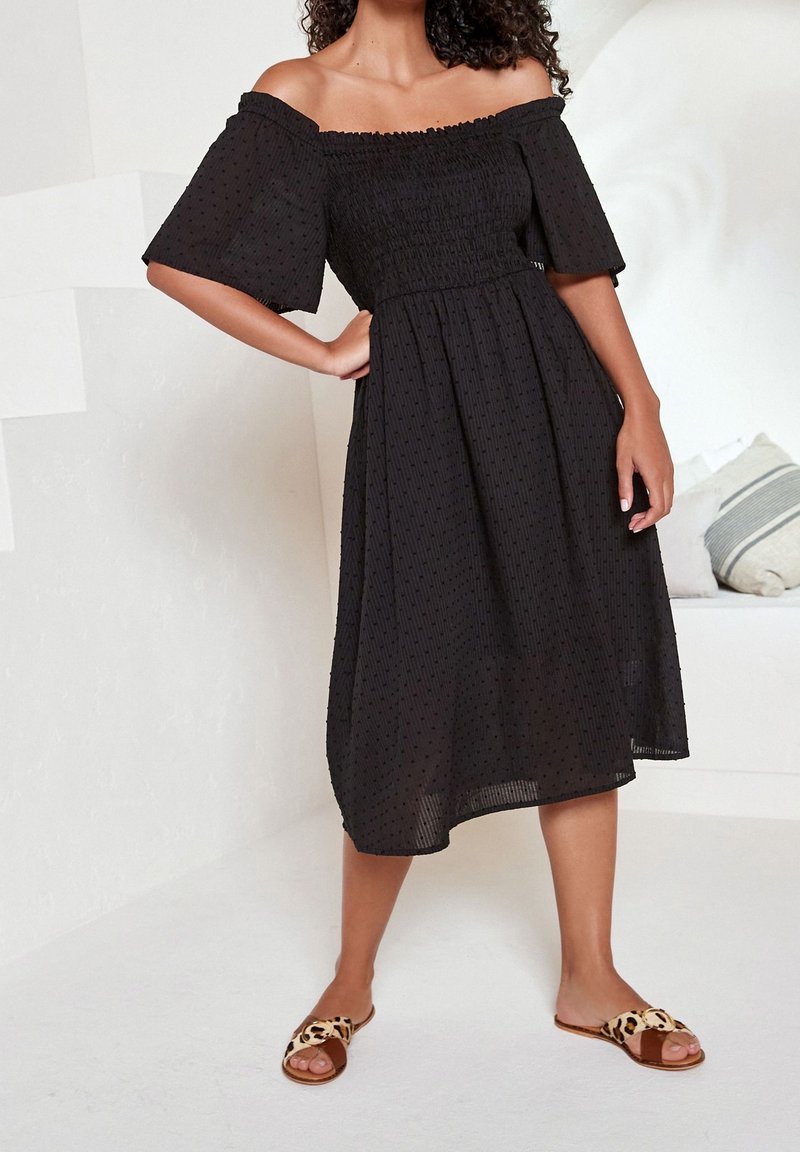 Robe noire épaules dénudées avec un corsage smocké, texture plissée et longueur mi-mollet. Portée avec des sandales à enfiler imprimé léopard.