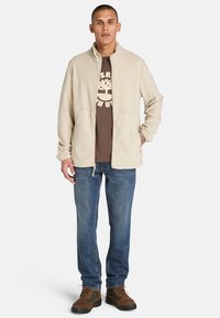Beige fleecejacka med dragkedja framtill, hög krage och sidofickor, bärs över en brun t-shirt, ihop med blå jeans och bruna skor.