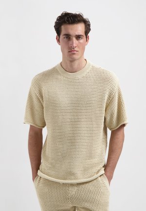 Gehaakte sweater met korte mouwen in lichtbeige, voorzien van een gestructureerd, open patroon en geribbelde halslijn en mouwen. Het model staat met de handen in de zakken.