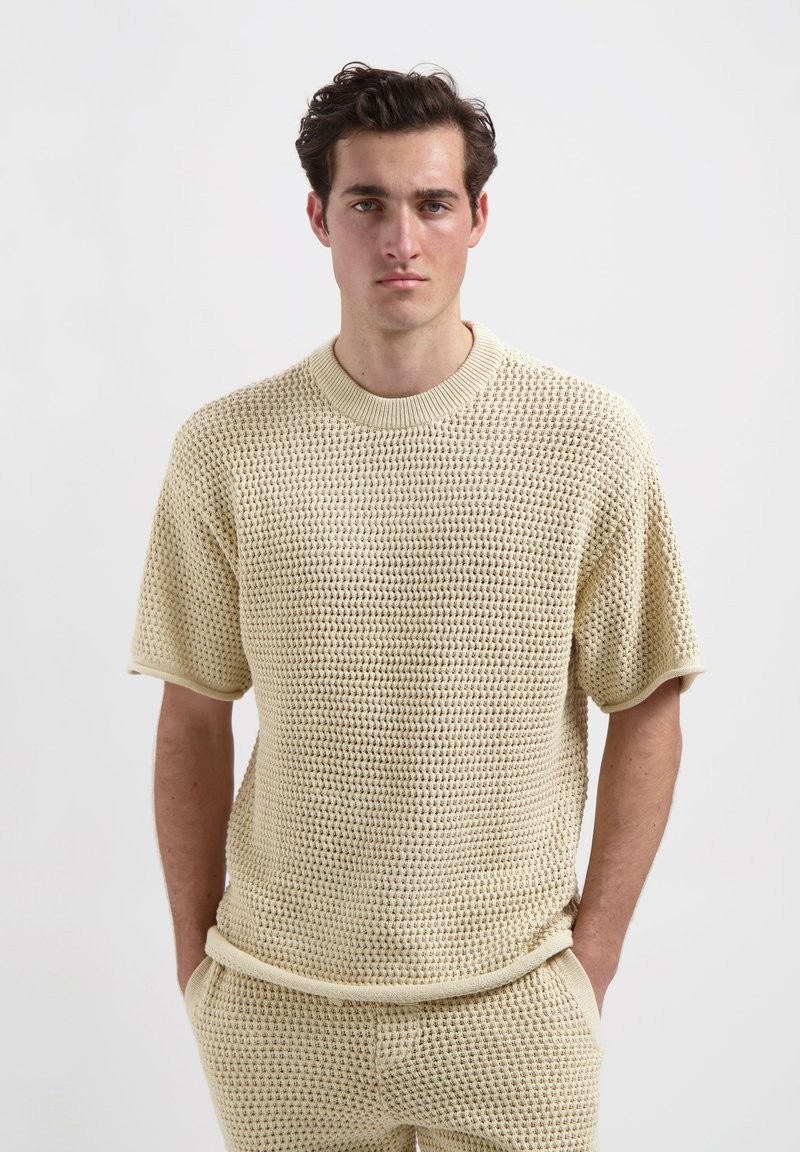 Gehaakte sweater met korte mouwen in lichtbeige, voorzien van een gestructureerd, open patroon en geribbelde halslijn en mouwen. Het model staat met de handen in de zakken.